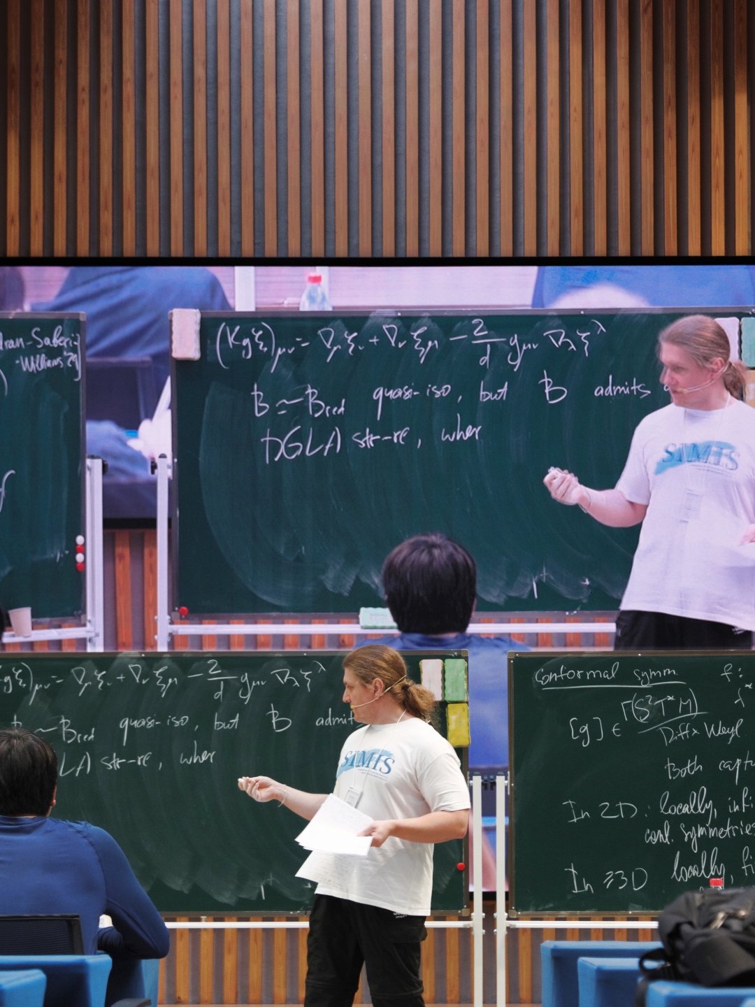 a lecturer giving a seminar infront of chalkboards with a live video projection in the background