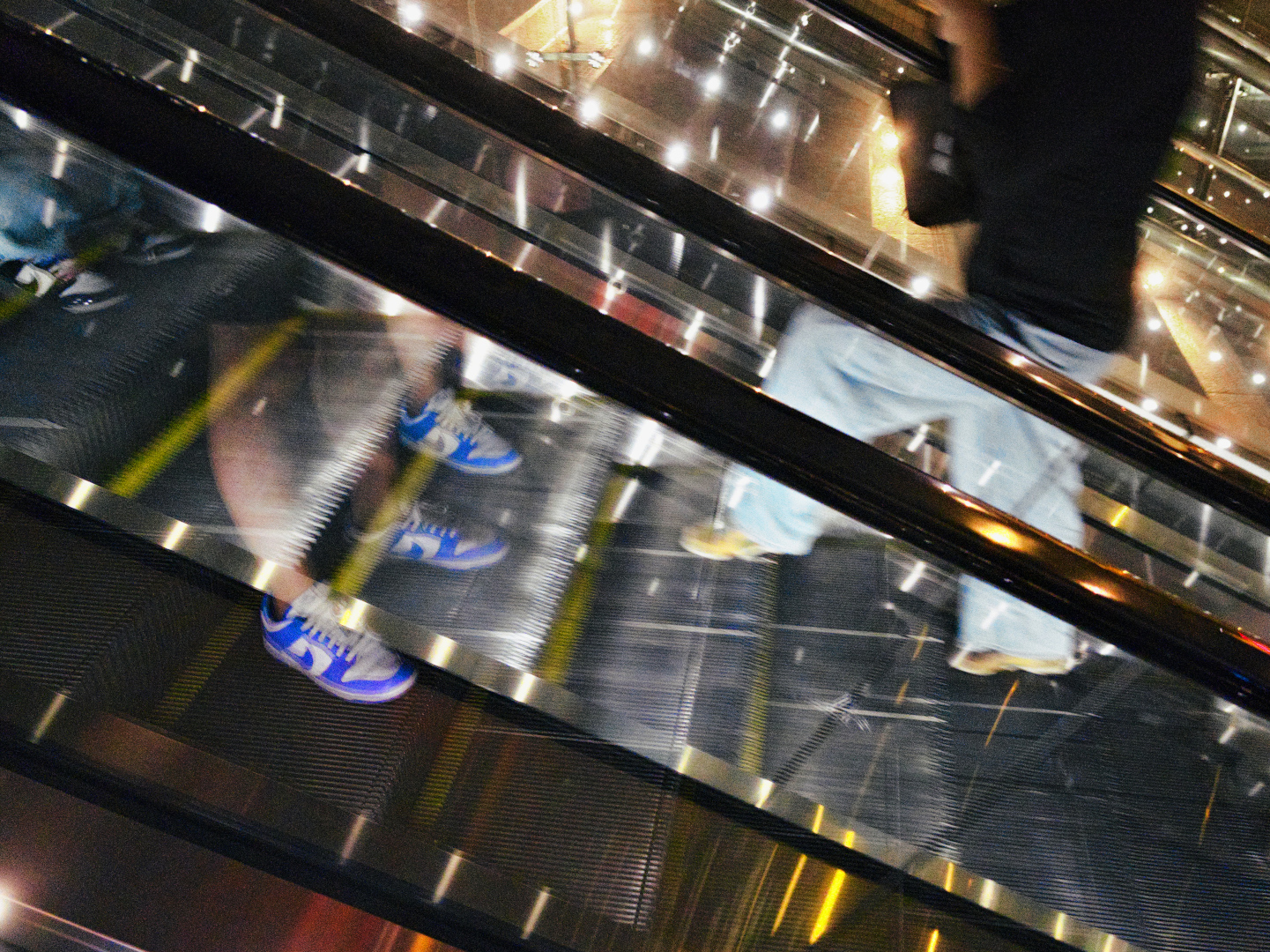 bottom halves of people going up and down an escalator at night with colorful lights